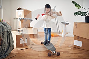 Little asian girl smiling, standing on boosted board, holding teddy bear and paint roller, surrounded with unpacked cardboard photo