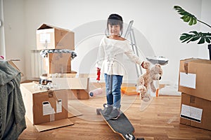 Little asian girl smiling, standing on boosted board, holding teddy bear and paint roller, surrounded with unpacked cardboard photo