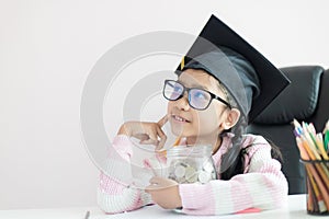 Little Asian girl wearing graduate hat hugging clear glass jar piggy bank and smile with happiness for money saving to wealthness
