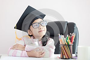 Little Asian girl wearing graduate hat hugging clear glass jar piggy bank and smile with happiness for money saving to wealthness