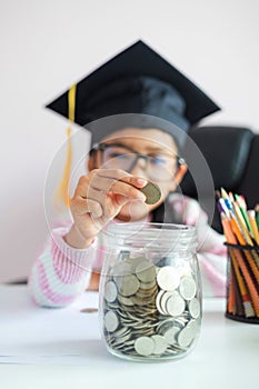 Little Asian girl wearing graduate hat putting the coin into clear glass jar piggy bank and smile with happiness for money saving