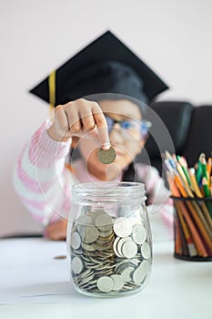 Little Asian girl wearing graduate hat putting the coin into clear glass jar piggy bank and smile with happiness for money saving