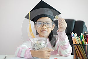 Little Asian girl wearing graduate hat putting the coin into clear glass jar piggy bank and smile with happiness for money saving