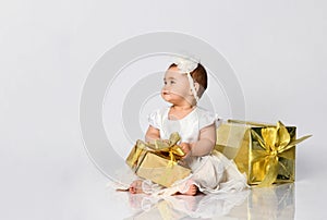 Little babe in dress and headband, barefoot. She posing with two golden gift boxes, sitting on floor  on white. Close up