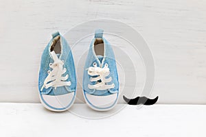 Little baby blue sneakers for boy and mustaches on a white wooden background