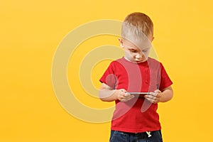 Little baby boy 3-4 years old in red t-shirt holding in hand, playing game on mobile phone isolated on yellow background