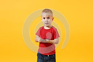 Little baby boy 3-4 years old in red t-shirt holding in hand, playing game on mobile phone isolated on yellow background