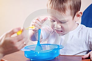 Little baby boy eating on a chair in the kitchen. Mom feeds holding in hand a spoon of porridge