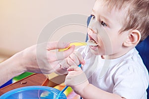 Little baby boy eating on a chair in the kitchen. Mom feeds holding in hand a spoon of porridge