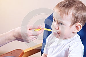 Little baby boy eating on a chair in the kitchen. Mom feeds holding in hand a spoon of porridge