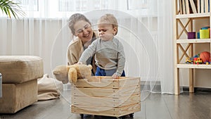 Little baby boy with mother collecting toys in wooden toy box at living room