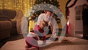 Little baby boy in Santa Claus costume crawling to his mother sitting under Christmas tree. Families and children