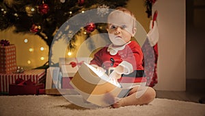 Little baby boy in Santa Claus costume sitting under Christmas tree and holding magical glowing Christmas present in