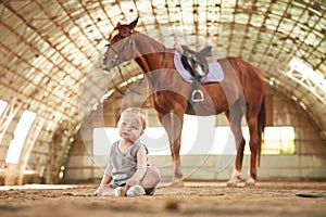 Little baby boy is sitting on the ground with horse indoors