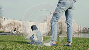 Little baby boy squatting plays on the grass sitting with his back to the camera and his young mother standing near him