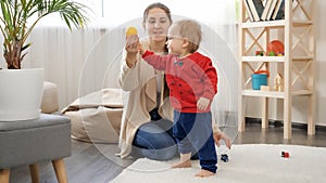 Little baby boy taking colorful toys from his mother and walking on soft carpet in living room. Baby development, family playing