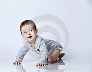 Little baby boy toddler in grey casual jumpsuit and barefoot crawling on floor, smiling and looking up over white wall background