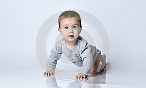 Little baby boy toddler in grey casual jumpsuit and barefoot crawling on floor, smiling and looking up over white wall background