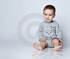 Little baby boy toddler in grey casual jumpsuit and barefoot sitting on floor and smiling over white wall background
