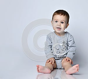 Little baby boy toddler in grey casual jumpsuit and barefoot sitting on floor and smiling over white wall background