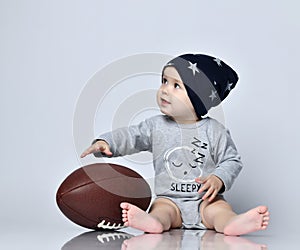 Little baby boy toddler in grey casual jumpsuit, black cap with stars and barefoot sitting on floor with rugby ball