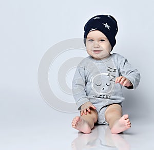 Little baby boy toddler in grey casual jumpsuit, black cap with stars and barefoot sitting on floor, smiling and gesticulating