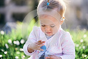 Little baby with a chamomile behind her ear sits on a green lawn and opens a plastic bottle. Close-up