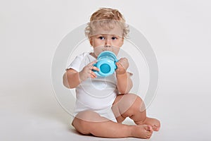 Little baby child drinking from blue bottle, cute child wearing bodysuit sitting on floor isolated over white background, blond