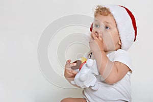 Little baby in christmas hat looking away with astonishing look, covering his mouth with tiny fingers, holding plastic dog toy,