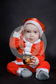 Little baby dressed as Santa Claus sits on a dark background and plays with Christmas toys