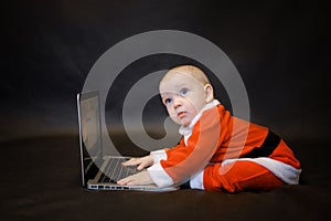 Little baby dressed as Santa Claus sits on a dark background and writes an email to Santa Claus on a laptop