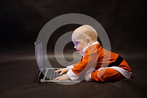 Little baby dressed as Santa Claus sits on a dark background and writes an email to Santa Claus on a laptop