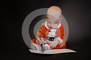 Little baby dressed as Santa Claus sits on a dark background and writes a letter to Santa Claus