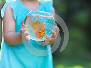 Little baby girl holding a fishbowl with a goldfish on a nature photo