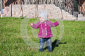 Little baby girl in jeans jacket and hat making learning to walk his first steps on the lawn in the green grass