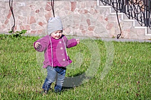 Little baby girl in jeans jacket and hat making learning to walk his first steps on the lawn in the green grass