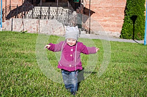 Little baby girl in jeans jacket and hat making learning to walk his first steps on the lawn in the green grass