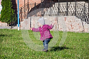 Little baby girl in jeans jacket and hat making learning to walk his first steps on the lawn in the green grass