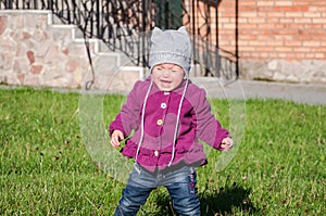 Little baby girl in jeans jacket and hat making learning to walk his first steps on the lawn in the green grass it has not very go