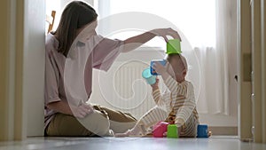 Little baby girl and mommy play with colorful toys at home sitting on floor. Mother and daughter laughing having fun