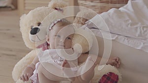 Little baby girl in a pink dress and diaper lying on the bear toy on the bed at home, raising feet up. Concept of a