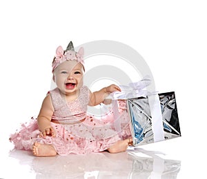 Little baby girl in pink princess dress, crown and shoes sitting on floor, laughing and holding present birthday box with bow