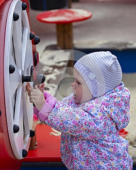 Little baby girl playing on the playground. One-year-old child in a hat and jacket plays with children`s carousels and sports