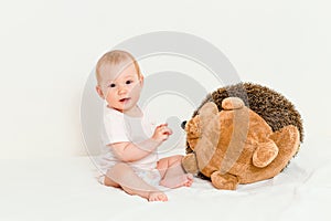 Little baby girl plays with a big soft hedgehog toy sitting on the bed on white background