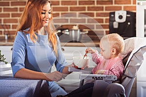 Little baby girl sits in high chair and feeding with spoon her beautiful mother