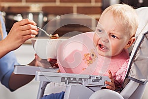 Little baby girl sits in high chair and feeding with spoon her beautiful mother