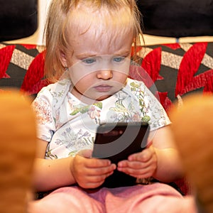 Little baby girl sitting on sofa and holding smarthphone. Mobile addiction