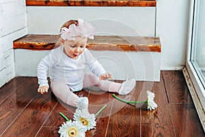 Little baby girl wearing spring wreath siting on floor in bright light living room near window and playing with gerbera