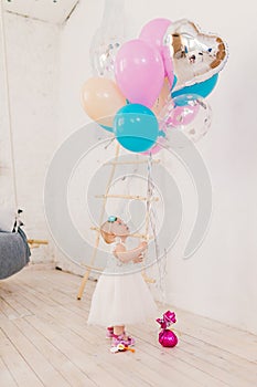 A little baby girl in white lavish dress is standing in living room in the house near the decorative staircase and