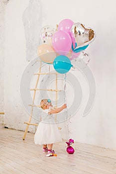 A little baby girl in white lavish dress is standing in living room in the house near the decorative staircase and